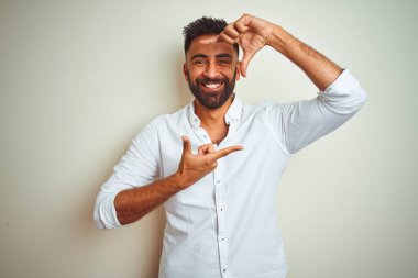 Zarif bir gömlek giyen genç Hintli adam izole edilmiş beyaz arka planda duruyor gülümsüyor ve elleri ve parmakları gülen yüzlü bir çerçeve yapıyor. Yaratıcılık ve fotoğrafçılık kavramı.