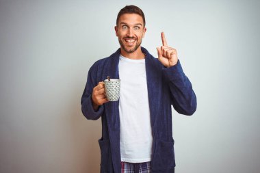 Rahat pijama ve bornoz giyen bir adam izole arka planda bir fincan kahve içiyor. Elle ve parmakla göstererek çok mutlu.
