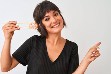 Genç ve güzel bir kadın yemek çubuklarıyla suşi yiyor. İzole edilmiş beyaz arka planda duruyor. Elleri ve parmakları kenarı işaret ederek çok mutlu.