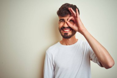 Dövmeli, tişörtlü genç adam izole edilmiş beyaz arka plan üzerinde duruyor. El hareketleriyle gülümsüyor, gözleriyle mutlu yüzle parmaklarının arasından bakıyor..