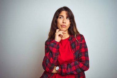 Kırmızı tişört ve ceket giyen genç ve güzel bir kadın beyaz izole edilmiş arka plan üzerinde duruyor ve elleri çenesinde soru sormayı düşünüyor, düşünceli bir ifade. Düşünceli bir yüzle gülümsüyor. Şüphe kavramı.
