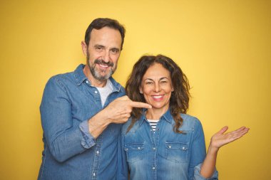 Güzel orta yaşlı çift, izole edilmiş sarı arka planda durarak hayretler içinde kameraya bakıp gülümserken elleri ve parmaklarıyla işaret ediyorlar..