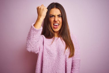 İzole edilmiş pembe arka planda dikilen sıradan bir süveter giyen genç ve güzel bir kadın öfkeyle bağırırken kızgın ve kızgın bir şekilde yumruğunu kaldırıyor. Öfke ve agresif kavram.