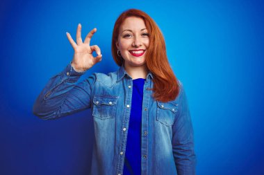 Genç, güzel, kızıl saçlı, kot gömlek giyen, mavi izole edilmiş arka plan üzerinde duran, gülümseyen, el ve parmaklarıyla 