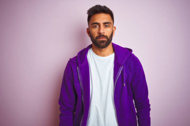 Mor eşofman giyen genç Hintli adam izole edilmiş pembe arka planda duruyor. Yüzünde ciddi bir ifadeyle rahatlamış. Basit ve doğal bir şekilde kameraya bakıyorsun..