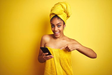 Duş üstüne havlu giyen Afro kadın izole edilmiş sarı arka plan üzerinde smatrphone kullanıyor. Elle ve parmakla işaret ederek çok mutlu.