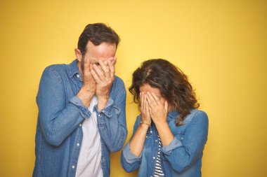 Güzel bir orta yaşlı çift, izole edilmiş sarı arka plan üzerinde kot gömlek giyiyorlar ve ağlarken yüzlerini ellerle kaplayan üzgün bir ifade var. Depresyon kavramı.