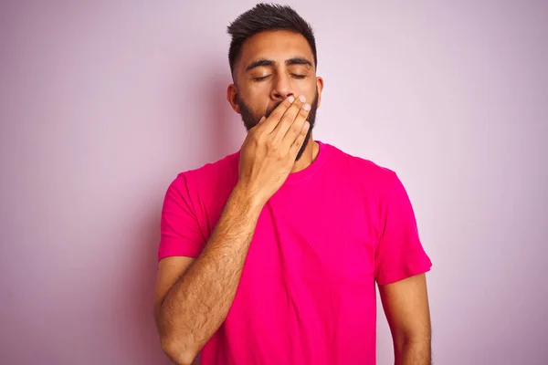 Young indian man wearing t-shirt standing over isolated pink background bored yawning tired covering mouth with hand. Restless and sleepiness.