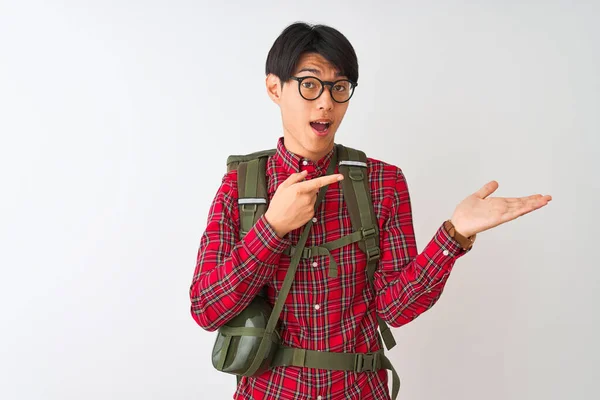 Chinese hiker man wearing backpack canteen glasses over isolated white background amazed and smiling to the camera while presenting with hand and pointing with finger.