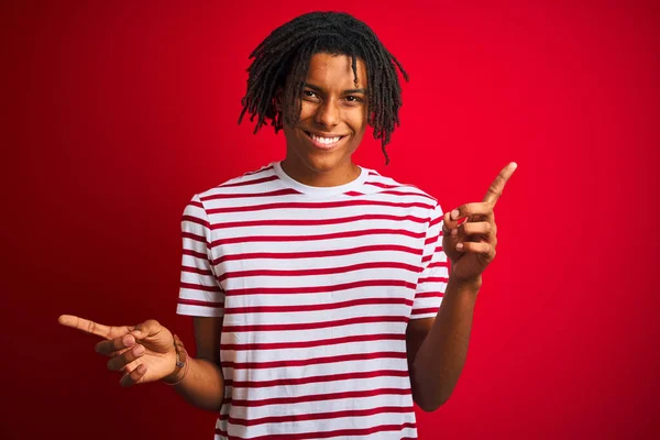 穿着条纹T恤的年轻黑人男子站在孤立的红色背景上 自信地微笑着 用手指指向不同的方向 广告的复制空间 — 图库照片