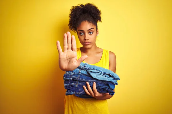 Young african american woman holding stack of jeans over isolated yellow background with open hand doing stop sign with serious and confident expression, defense gesture