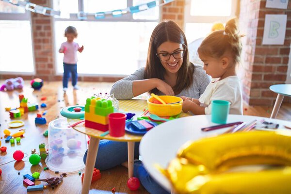 Young beautiful teacher and toddlers playing on the table with lots of toys at kindergarten