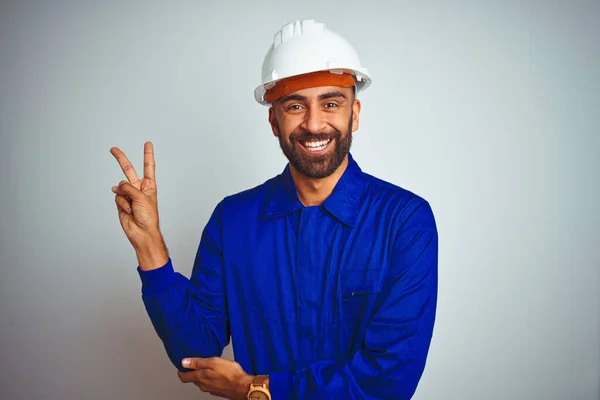 勝利の印をしているカメラで幸せな顔のウィンクで微笑む隔離された白い背景の上に制服とヘルメットを身に着けているハンサムなインドの労働者の男 第二番 — ストック写真