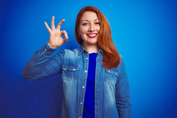 青い隔離された背景の上に立つデニムシャツを身に着けている若い美しい赤毛の女性は手と指でOkサインをして肯定的な笑顔 成功した表現 — ストック写真