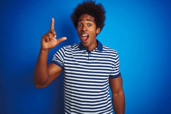 African american man with afro hair wearing striped polo standing over isolated blue background pointing finger up with successful idea. Exited and happy. Number one.