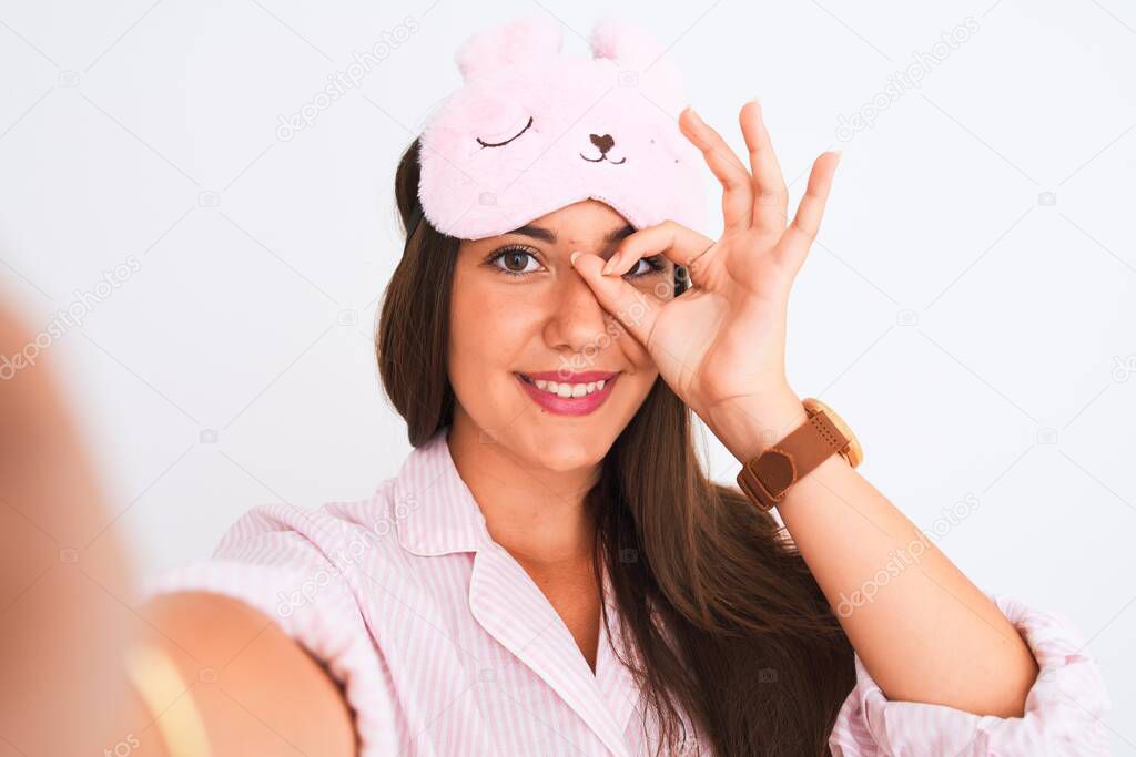 Girl wearing pajama and sleep mask make selfie by camera over isolated white background with happy face smiling doing ok sign with hand on eye looking through fingers