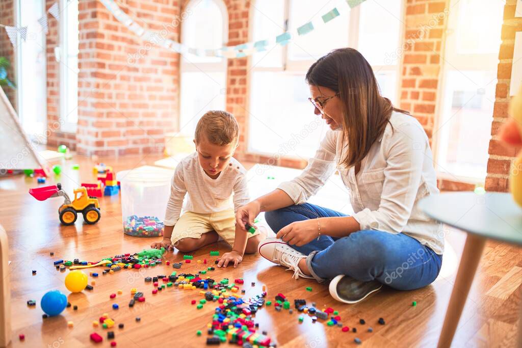 Beautiful teacher and toddler playing with building blocks around lots of toys at kindergarten