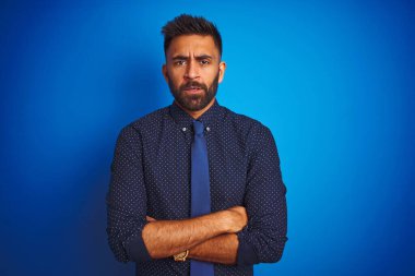 Genç Hintli iş adamı zarif bir gömlek ve kravat giyerek izole edilmiş mavi arka plan üzerinde duruyor şüpheci ve gergin, yüzünde çapraz kollarla onaylamayan bir ifade. Negatif kişi.