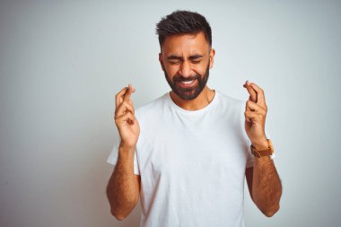 İzole edilmiş beyaz arka planda duran tişört giyen genç Hintli adam umut ve gözler kapalı bir şekilde gülümseyerek işaret ediyor. Şans ve batıl inanç..