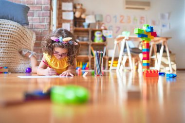 Anaokulunda kağıt ve kalem kullanarak yere uzanmış, gözlüklü, tek boynuzlu at diademi takan güzel bir çocuk.