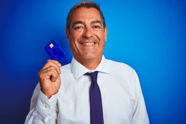 Yakışıklı orta yaşlı bir işadamı, mavi arka planda kredi kartı tutuyor. Mutlu bir yüzü var, gülümsüyor ve kendine güvenen bir gülümsemesi var.