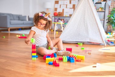 Gözlük takan ve tek boynuzlu at diademi takan güzel çocuk anaokulunda yapı taşlarıyla oynuyor.