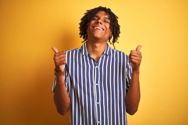 Rastgele çizgili tişört giyen Afro bir adam izole edilmiş sarı arka plan başarısı tabelasının üzerine el hareketleriyle olumlu jest yapıyor, gülümsüyor ve mutlu. Neşeli ifade ve kazanan jest.