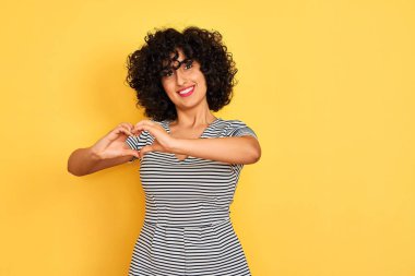 Sarı arka planda çizgili elbise giyen kıvırcık saçlı genç Arap kadın kalp sembolü ve elleri ile şekil veren aşkla gülümsüyor. Romantik konsept.