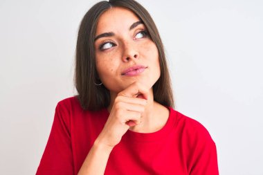 Kırmızı günlük tişört giyen genç güzel bir kadın izole edilmiş beyaz arka planda dikiliyor. Ciddi bir yüz ifadesi var. Soru sormayı düşünüyor.