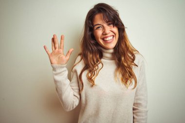 Kışlık süveter giyen genç bir kadın izole edilmiş beyaz arka plan üzerinde duruyor ve beş numaralı parmaklarıyla işaret ederken kendinden emin ve mutlu bir şekilde gülümsüyor..