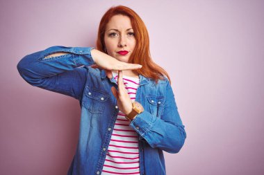 Güzel kızıl saçlı kadın kot gömlek ve çizgili tişört giyip pembe arka planda takılıyor. Elleriyle jest yapıyor, hayal kırıklığına uğramış ve ciddi bir yüz ifadesi takınıyor.