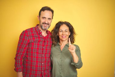 Izole sarı arka plan üzerinde Güzel orta yaş çift gösteren ve emin ve mutlu gülümseyerek parmak sayısı bir ile işaret.