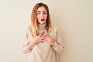 Şık balıkçı yaka kazak giyen kızıl saçlı kadın izole edilmiş beyaz arka plan üzerinde duruyor elleri göğsünde, gözleri kapalı ve yüzünde minnet dolu bir jest ile gülümsüyor. Sağlık kavramı.