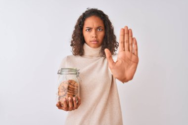 Genç Brezilyalı bir kadın elinde bir kavanoz kurabiyeyle izole edilmiş beyaz arka planda duruyor. Açık elle dur işareti yapıyor. Ciddi ve kendinden emin bir ifadeyle.