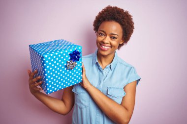 Afro-Amerikalı bir kadın doğum günü hediyesini pembe izole edilmiş arka planda tutarken mutlu bir yüzle ayakta duruyor ve gülümsüyor dişlerini gösteren bir gülümsemeyle.