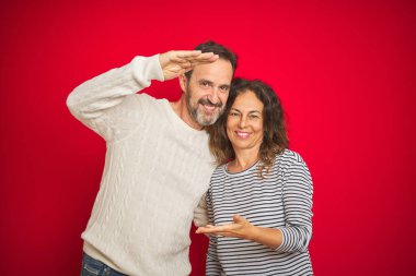 Güzel bir orta yaşlı çift, izole edilmiş kırmızı arka plan jestleri üzerine kışlık süveter giyiyorlar. Ellerinde büyük ve büyük boy işareti, ölçü sembolü var. Kameraya bakıp gülümsüyor. Ölçüm konsepti.