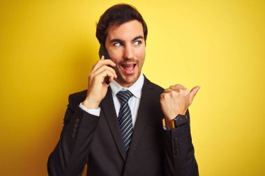 Akıllı telefonda konuşan genç yakışıklı işadamı izole edilmiş sarı arka planda işaret ediyor ve gülümseyen bir yüzle baş parmağıyla onu gösteriyor.