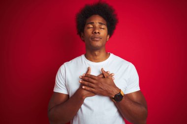 Afro saçlı, beyaz tişörtlü, izole edilmiş kırmızı arka planda duran, elleri göğsünde, gözleri kapalı ve yüzünde minnet dolu bir jest olan genç bir Amerikalı. Sağlık kavramı.