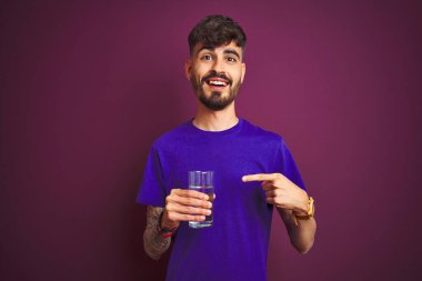 Dövmeli genç adam izole edilmiş mor arka planda duran su bardağını içerken mutlu bir şekilde elini ve parmağını gösterdi.