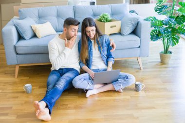 Genç çift bilgisayar laptop kullanarak yeni ev katında oturan ve yeni bir eve taşımak için mutlu gülümseyen