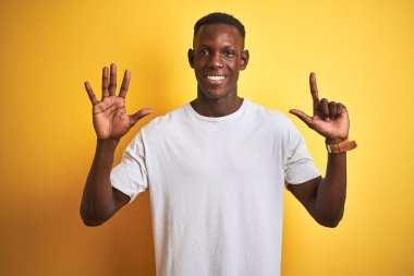 Beyaz tişörtlü genç Afro-Amerikalı adam izole edilmiş sarı arka planda duruyor ve yedinci parmaklarını gösterirken kendinden emin ve mutlu bir şekilde gülümsüyor..