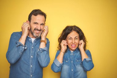 Güzel orta yaşlı çift, izole edilmiş sarı arka planda dikiliyorlar. Yüksek sesli müzik seslerini bastırmak için parmaklarıyla kulaklarını kapatıyorlar. Sağır kavramı.
