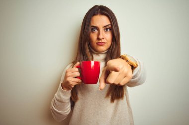 Genç ve güzel bir kadın izole edilmiş beyaz arka planda dikilip kırmızı kahve içerek kamerayı işaret ediyor ve sen, el işareti, ön taraftan pozitif ve kendinden emin bir hareket.