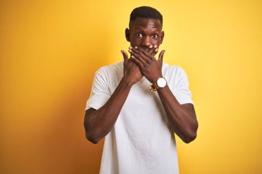 Beyaz t-shirt giyen genç Afro-Amerikalı adam izole edilmiş sarı arka planda dikiliyor. Ağzını yanlışlıkla ellerle kapatıyor. Gizli konsept.