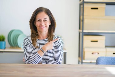 Evde masada oturan orta yaş yaşlı kadın mutlu ve doğal ifade ile yan yana el ve parmak ile işaret yüzünde bir gülümseme ile neşeli