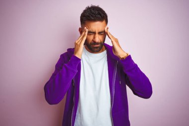 Mor eşofman üstü giyen genç Hintli adam stres yüzünden kafa üstü başı ağrıyan pembe arka planda dikiliyor. Migren ağrısı..