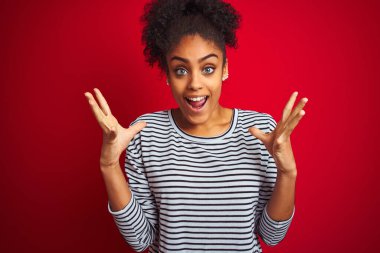 Siyah çizgili tişört giyen Afro-Amerikalı bir kadın izole edilmiş kırmızı arka planda durmuş çılgın ve başarıyı kutlarken kollarını kaldırmış ve gözleri açık bir şekilde heyecanla haykırıyordu. Kazanan konsept