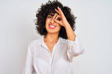 Kıvırcık saçlı genç bir Arap kadın izole edilmiş beyaz arka planda günlük gömlek giyiyor. El hareketleriyle gülümsüyor ve mutlu yüzüyle parmaklarının arasından bakıyor..