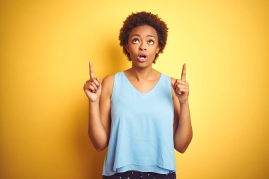 Güzel afro-amerikan kadın zarif bir gömlek giyerek izole edilmiş sarı arka planda şaşkın şaşkın yukarı bakıyor ve parmaklarıyla işaret ediyor ve kollarını kaldırıyor..