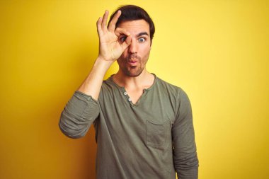 Sarı arka plan üzerinde sıradan tişörtler giyen genç yakışıklı adam şaşırmış yüz ifadesiyle şaşırmış, parmaklarının arasından bakan bir jest yapıyor. İnançsız ifade.